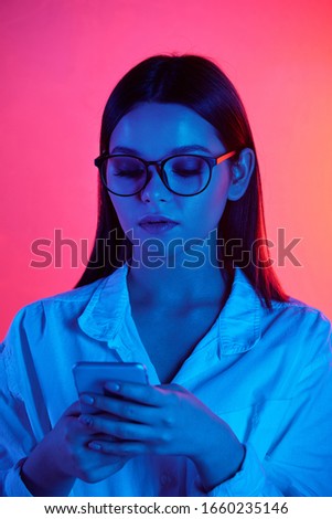 Serious female office worker in eyeglasses and white shirt using mobile phone in isolation on pink neon background
