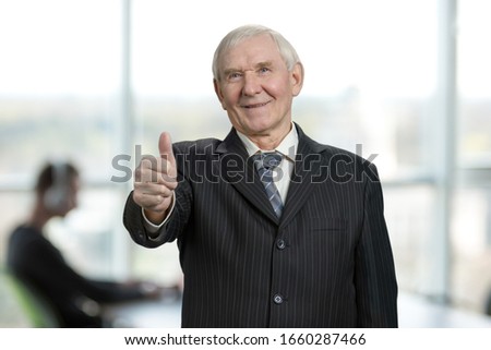 Cheerful business man with thumb up. Old senior male in suit in right office background.