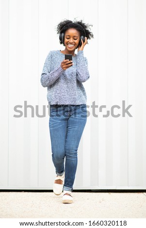 Full length portrait happy young black woman walking listening to music with cellphone and headphones by white wall