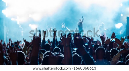 Rear back view of audience crowd people fans raising hands enjoying live music festival concert event concept shooting on phones rock band silhouettes performance sing on night club outdoor stage.