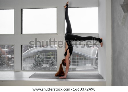 Young skinny woman doing yoga handstand against window