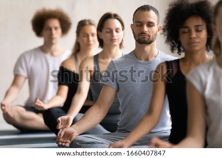 Group of diverse people meditating together visualizing during yoga morning session, focus on Caucasian man seated cross-legged in row with associates, no stress, spiritual practise, lifestyle concept Royalty-Free Stock Photo #1660407847