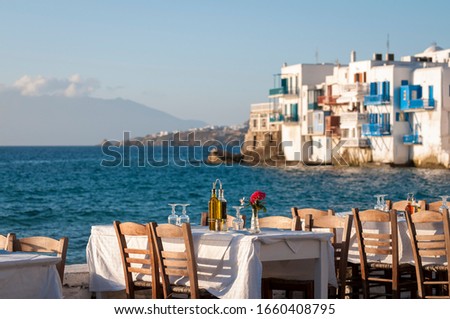 Bright scenic afternoon view of empty tables waiting for sunset diners set up along the harbor in the old town of Mykonos, Greece Royalty-Free Stock Photo #1660408795