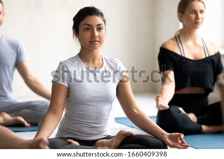 Indian woman with Caucasian associates in sportswear working out meditating seated cross-legged on carpets during hatha yoga class, wellness, spiritual practice for enhancing of self-awareness concept