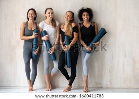 Full length of multi-ethnic vivacious women smiling looking at camera holding carpets leaned on wall background, comfy tops and pants sportswear advertisement, sport club instructors portrait concept Royalty-Free Stock Photo #1660411783