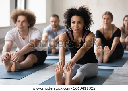 Wearing comfy stylish grey black color activewear group of people working out performing Seated Forward Bend exercise. Mixed-race woman and associates gather together for yoga training at sport club Royalty-Free Stock Photo #1660411822