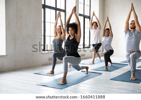 Young diverse people with African ethnicity yoga instructor standing on mats performing Warrior one asana or Virabhadrasana 1 pose. Work out physical activity in modern studio class, wellness concept Royalty-Free Stock Photo #1660411888