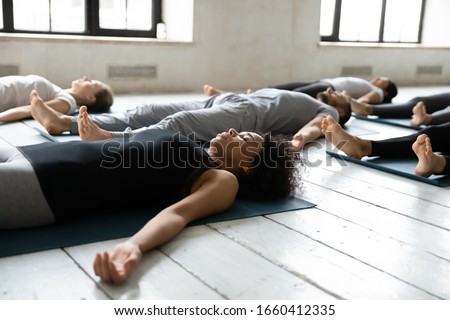 Diverse people resting after work out lying on carpets performing Shavasana or Corpse Dead position focus on African female, final pose of yoga class deep restoration, body and mind relaxation concept Royalty-Free Stock Photo #1660412335