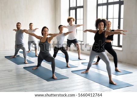 Multinational millennial girls and guys standing barefoot on mats performing Warrior II or Virabhadrasana position. Session led by mixed-race woman trainer showing exercise to people during yoga class Royalty-Free Stock Photo #1660412764