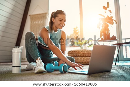 A sporty woman in sportswear is sitting on the floor with dumbbells and a protein shake or a bottle of water and is using a laptop at home in the living room. Sport and recreation concept. Royalty-Free Stock Photo #1660419805