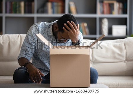 Stressed young multiracial man sitting on couch at home, feeling desperate about ordering wrong item. Depressed african american male client unpacked delivery box, dissatisfied with purchase. Royalty-Free Stock Photo #1660489795