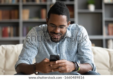 Smiling young african american man in glasses sitting on cozy couch, typing message on smartphone for friends or beloved woman girlfriend. Happy biracial guy playing online game on mobile phone.