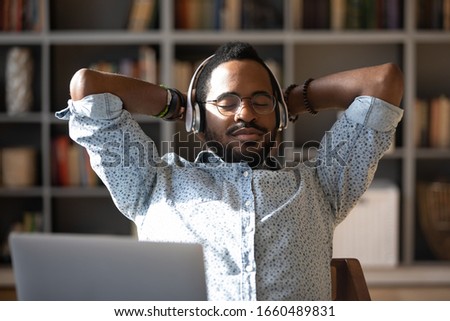 Head shot peaceful young african american businessman folded arms behind head, listening to favorite classic music in headphones, relaxing enjoying break pause time with closed eyes alone in office.
