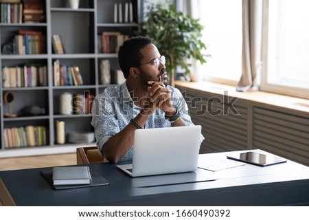 Thoughtful mixed race businessman sitting at table with computer, looking away. Distracted from study job young african american man lost in thoughts, thinking of difficult decision at home office. Royalty-Free Stock Photo #1660490392