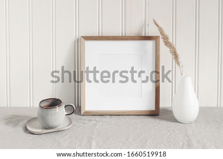 Square empty wooden frame mockup with modern ceramic vase, dry grass, cup of coffee on table. White beadboard wainscot wall paneling background. Scandinavian interior, home design. Art concept.