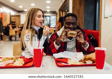 Two young friends sit at cafe, drink cola, eat burgers and fast food.