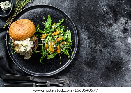 Homemade cheeseburger with blue cheese, bacon, marbled beef and onion marmalade, a side dish of salad with arugula and oranges. Black background. Top view. Copy space