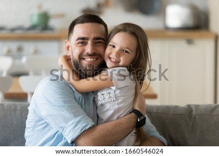 Portrait of smiling young father hug cuddle cute little preschooler daughter look at camera posing at home together, happy dad embrace small girl child show love relax on leisure domestic weekend