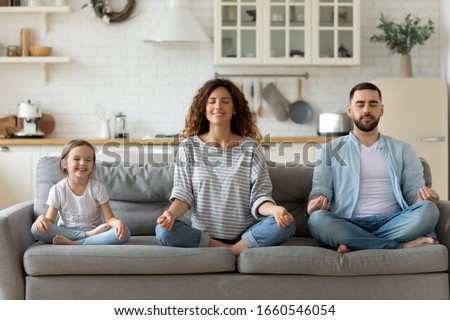 Calm young family with little daughter sit on couch practice yoga together, happy parents with small preschooler girl child rest on sofa meditate relieve negative emotions on weekend at home Royalty-Free Stock Photo #1660546054
