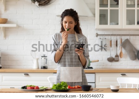 Smiling young woman in apron stand in modern kitchen preparing salad use smartphone read recipe, happy millennial female cooking making dinner or lunch chatting browsing on modern cellphone