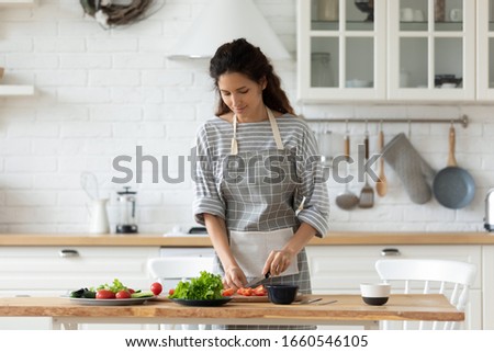 Millennial woman in apron stand at kitchen table chop vegetables preparing salad for dinner or lunch, young housewife cooking healthy tasty breakfast at home, dieting, vegetarian concept Royalty-Free Stock Photo #1660546105