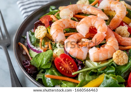 Close up of fresh salad with shrimp, tomato, carrots, onions, peppers and mixed greens (arugula, spring mix) on marble background with fork and striped napkin. Healthy food. Clean eating. Keto.