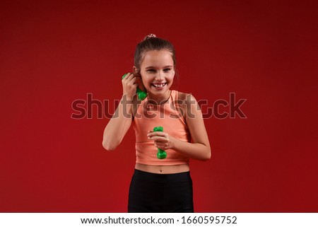 A cute kid, girl is engaged in sport, she is smiling at camera while exercising with dumbbells. Isolated on red background. Fitness, training, active lifestyle concept. Horizontal shot