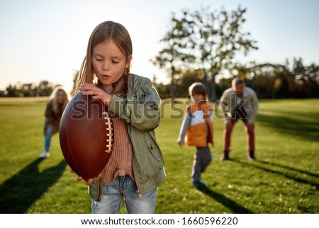 Portrait of small girl holding an oval brown leather rugby ball while playing american football with her parents and brother in park. Family and kids, nature concept. Horizontal shot