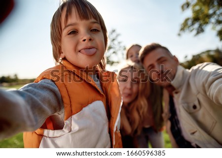 Portrait of cheerful little boy taking pictures with his family using smartphone in the park on a sunny day. Family, parenthood, and leisure concept. Horizontal shot. Front view