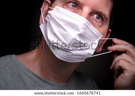 Close up of a caucasian man putting on a medical face mask with one hand stretching the elastic band and putting the protective mouthpiece on Royalty-Free Stock Photo #1660678741