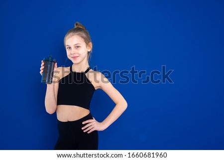 a little girl in a sports uniform on a pheolete yoga Mat does gymnastics on a blue background and drinks water from a drinking bowl