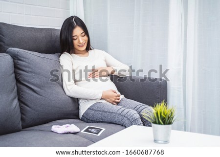 beautiful asian pregnant woman placing hands on baby lump feeling heartbeat smiling joyfully, sitting on sofa relaxing resting from tiredness, in living room with brick texture wall and white curtains