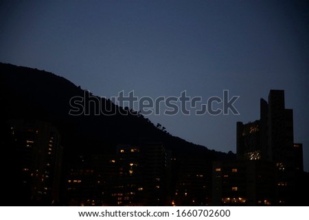 Night photo of high-rise buildings against the background of a mountain. Illuminated windows in darkness. Nature and modern architecture under deep blue sky.