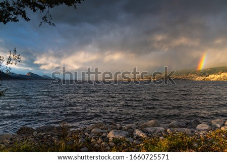 Dramatic storm clouds and rain passing over Okanagan Lake with view of a double rainbow above Naramata