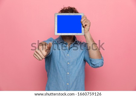 casual young guy hiding behind tab and making thumbs us sign, standing on pink background