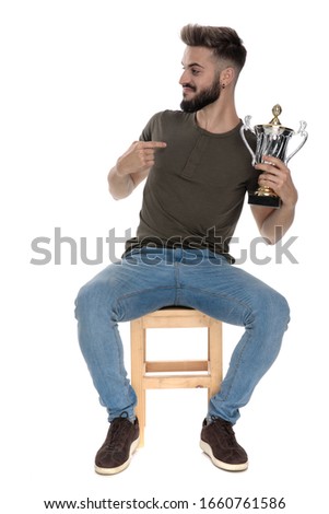 Happy casual man holding a trophy and pointing at it while sitting on a chair on white studio background
