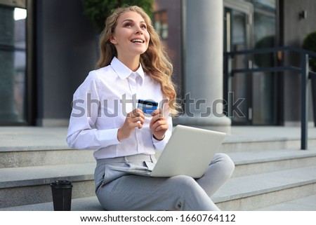 Online shopping concept. Young blonde woman holding a credit card and doing online payment with laptop outdoors.
