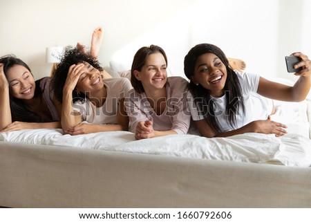 Beautiful African American girl taking selfie on phone with diverse girlfriends at sleepover or hen-party, four pretty young women wearing pajamas lying in bed, posing for photo together