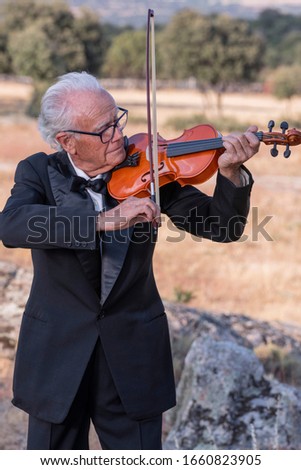 Elderly man, dressed in tuxedo, plays the violin in a natural environment
