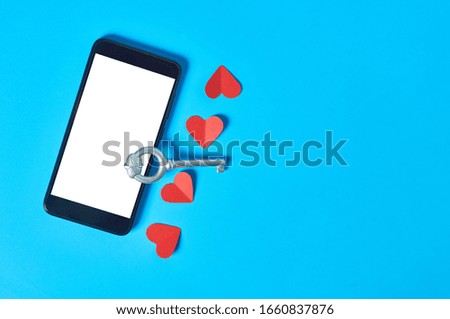 Black smartphone with isolated white screen for text, picture, photo and other graphics near paper hearts and vintage key on blue table. Secrets or forbidden love concept. Space for text. Top view