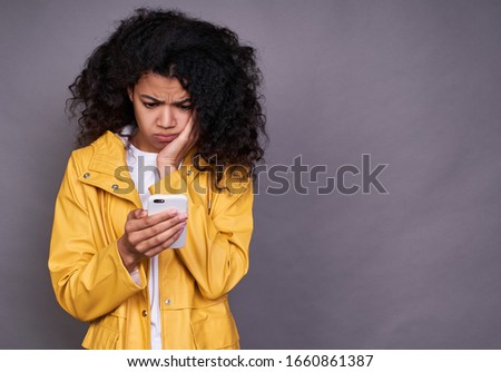 Pensive young cute pretty African American woman, with curly puffy hair, in a yellow raincoat, looking at a mobile phone with a sad expression on a gray background.
