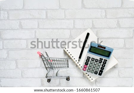 Top view objects , calculator, black pen on  notebook  with small mini shopping cart on  white gray brick background. Idea business and finance concept. sensitive focus.