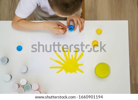 The child draws at a white table. The boy painted the sun and took the blue paint.