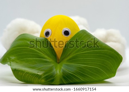 Children's craft egg painted with yellow paint. Eyes on the egg. An egg is hiding behind a green leaf. Close-up. Selective focus