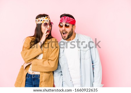 Young caucasian couple wearing a music festival clothes isolated is saying a secret hot braking news and looking aside
