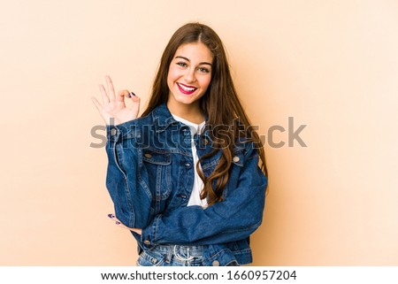 Young caucasian woman isolated en beige background winks an eye and holds an okay gesture with hand.