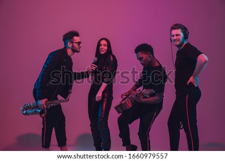 Group of young multiethnic musicians created band, dancing in neon light on pink background. Concept of music, hobby, festival, wellness. Joyful party host, dancer, singer, guitarist, saxophonist.