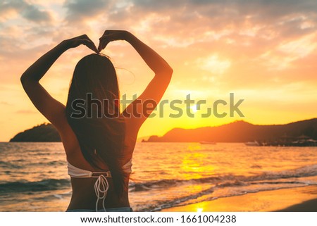 Summer travel vacation concept, Happy traveler asian woman with bikini relax and making hand in heart shape on sea beach at sunset