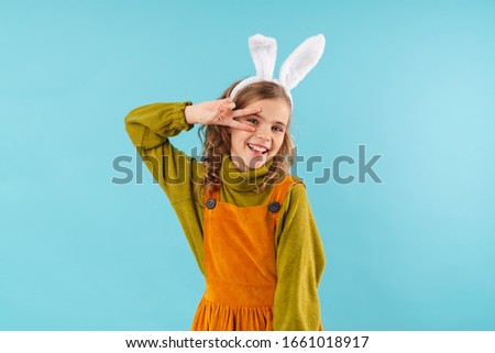 Photo of joyful curly girl in toy rabbit ears smiling and gesturing peace sign isolated over blue background