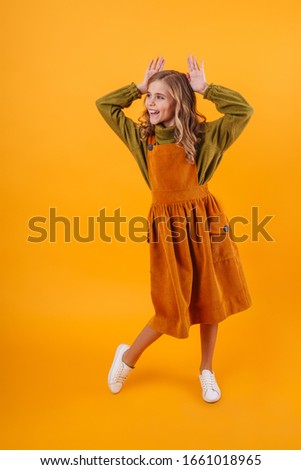 Photo of joyful curly girl showing rabbit ears behind her head and smiling isolated over blue background